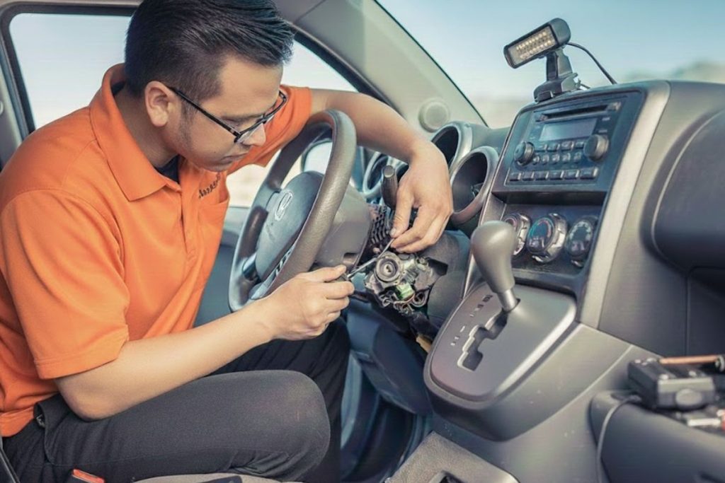 Locksmith wearing an orange shirt repairs a car ignition and steering column inside a vehicle, using tools near the dashboard.